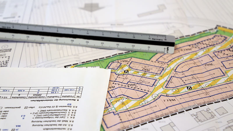 Das Foto zeigt die Planzeichnung eines Bebauungsplans, auf dem ein Textdokument und ein Maßstab liegen.