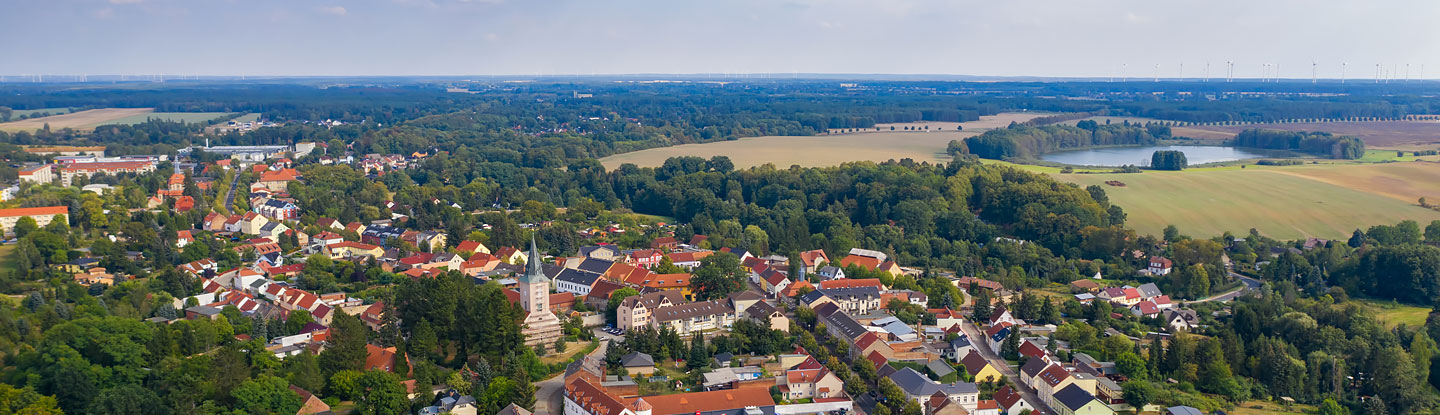 Blick von oben auf die Stadt Biesenthal. Im Vordergrund des Bildes befindet sich der Ortskern mit Kirche. Im Bildhintergrund befinden sich bewaldete sowie landwirtschaftlich genutzte Flächen. Auf der rechten oberen Seite des Bildes befindet sich zwischen Ackerflächen ein See.